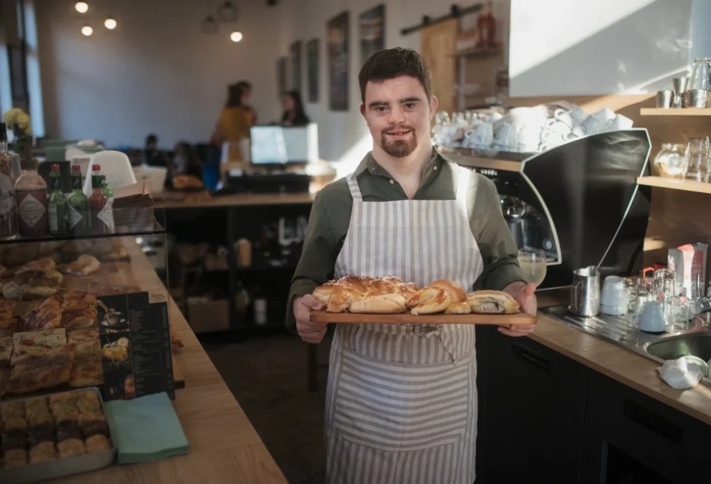 Disabled man with down syndrome working in cafe, holding plate of pastries